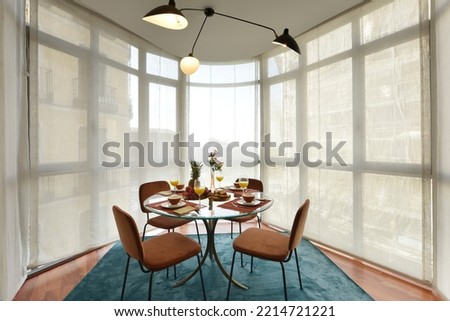 Beautiful circular beveled edge clear glass dining table with crockery of fresh fruit and sweet desserts, brown velvet upholstered chairs and bay window with blinds