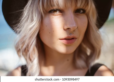Beautiful close up portrait of young blonde woman with piercing wearing black hat looking at the camera outdoors