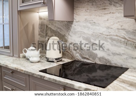beautiful closeup of custom designed kitchen, with marble looking quartz countertop and backsplash. cream electric kettle with porcelain tea accessories on the marble countertop next to ceramic hob