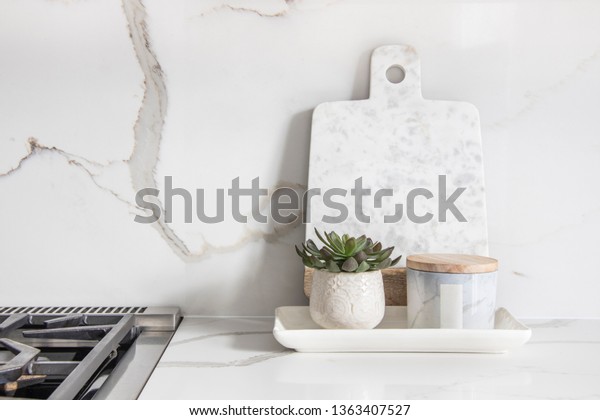 A beautiful closeup of a\
custom designed kitchen, with marble looking quartz countertop and\
backsplash. Decorated by marble cheese board and little indoor\
planter.