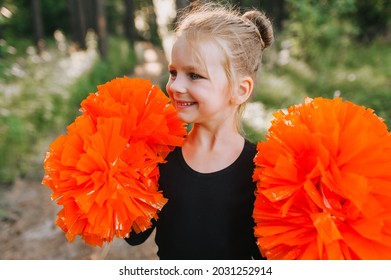 Beautiful Close-up Portrait Of A Smiling Blonde Girl, Child Cheerleader In A Black Suit With Orange Big Pom-poms In Her Hands On Nature In The Forest At Sunset. Sports Training For Cheerleading.