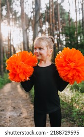 Beautiful Close-up Portrait Of A Smiling Blonde Girl, Child Cheerleader In A Black Suit With Orange Big Pom-poms In Her Hands On Nature In The Forest. Sports Training For Cheerleading.