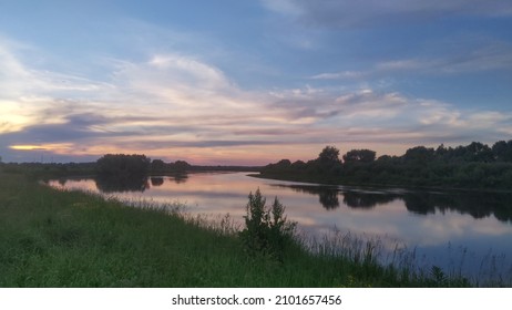 Beautiful cloudy sky at sunset over the river. The high bank of the river is overgrown with grass. The opposite bank of the river is low and there is a forest growing on it. - Powered by Shutterstock