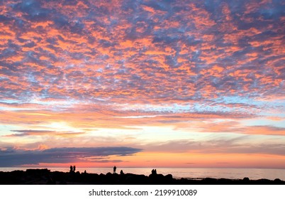 Beautiful Coastal New England Sunset. Dark Outlines (silhouettes) Of Unrecognizable People Along Breakwater Rocks At Cape Cod Beach In Dennis, Massachusetts Viewing Colorful Sky With Sunlit Clouds.