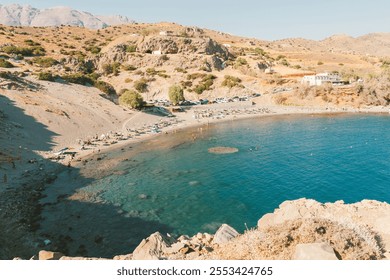 A beautiful coastal scene with a crescent-shaped beach curving into a calm turquoise sea. The beach is lined with sunbathers and umbrellas, while a few people are enjoying the water. - Powered by Shutterstock