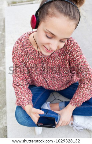 Beautiful college student girl sitting on bench using smart phone and headphones to listen to music, smiling outdoors. Teenager using technology, sensory perception leisure recreation lifestyle.