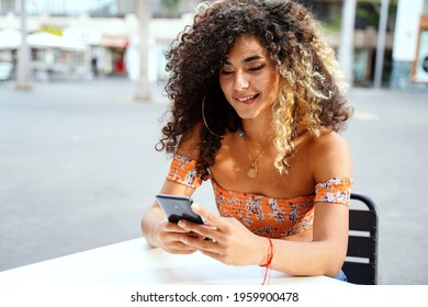 Beautiful Colombian Woman With Afro Hairstyle Using Mobile Phone, Flirting With Someone Or Posting New Photo On Social Media. Outdoor Photo. Relax Time In Cafe. 