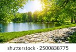 Beautiful and colorful spring summer nature scene with a lake in a park surrounded by green leaves of trees in the sunlight and a stone path in the foreground.