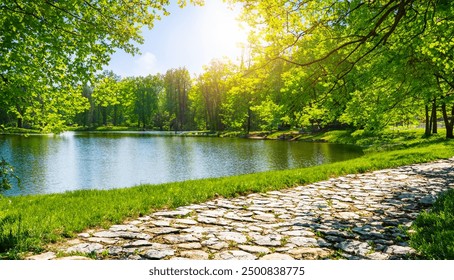 Beautiful and colorful spring summer nature scene with a lake in a park surrounded by green leaves of trees in the sunlight and a stone path in the foreground. - Powered by Shutterstock