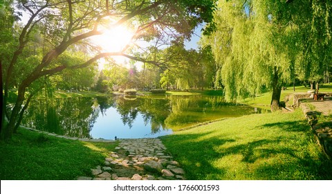 Красивый красочный летний весенний природный ландшафт с озером в парке, окруженный зеленой листвой деревьев в солнечном свете и каменной дорожкой на переднем плане.