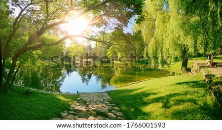 Beautiful colorful summer spring natural landscape with a lake in Park surrounded by green foliage of trees in sunlight and stone path in foreground.