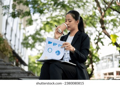 A beautiful and concentrated millennial Asian businesswoman is sipping coffee and examining business financial reports while sitting on the stairs outdoors. - Powered by Shutterstock