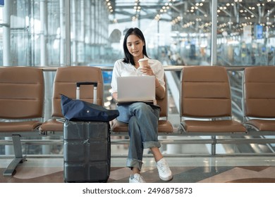 A beautiful, confident Asian businesswoman in casual clothes sits at a waiting seat in the airport, sipping coffee and working on her laptop computer, managing her work before her flight. - Powered by Shutterstock