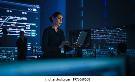 Beautiful Confident Female Project Manager works on Laptop Standing in Telecommunications System Control Room. In Background Big Screen Showing Infographics of Infrastructure, Charts, System Analysis