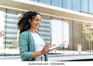 Beautiful confident successful latino or brazilian young woman with curly hair, business lady, financial expert, in elegant wear, standing outdoors with financial charts, analyze graphs, smiling - Powered by Shutterstock