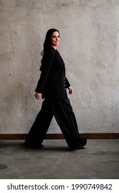 A Beautiful And Confident Woman In A Black Pantsuit Walks Along The Gray Wall. Women's Clothing.