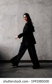 A Beautiful And Confident Woman In A Black Pantsuit Walks Along The Gray Wall. Women's Clothing.