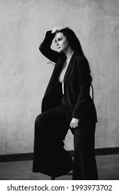 A Beautiful And Confident Woman In A Black Pantsuit On Naked Body On A Chair By The Gray Wall. Women's Clothing.