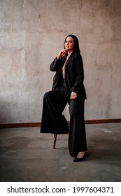 A Beautiful And Confident Woman In A Black Pantsuit On Naked Body On A Chair By The Gray Wall. Women's Clothing.