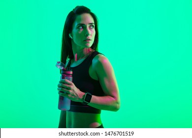 Beautiful Confident Young Fitness Girl Standing Isolated Over Green Neon Background, Wearing Wireless Earphones, Holding Water Bottle After Workout