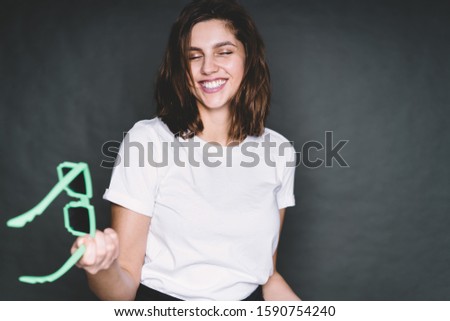 Beautiful contemporary brown shoulder length haired female with closed eyes standing in white t-shirt in studio and holding toy sunglasses mint color