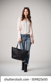 Beautiful Cool Woman Holding Black Shopping Bag And Walking On Grey Background. Fashion Girl Wearing Pink Cardigan, Jeans And Polka Dot Shirt. Stylish Casual Woman Holding A Big Bag And Looking Away.