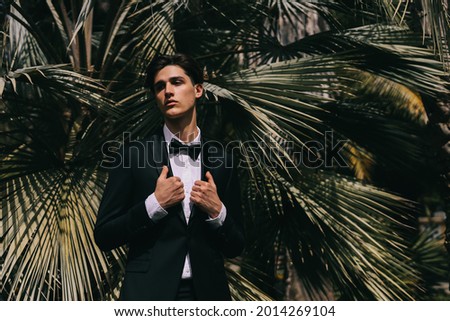 A beautiful cool young man in an elegant wedding suit poses and walks alone among plants and palm trees in an old park in nature outdoors, selective focus