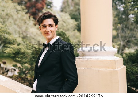 A beautiful cool young man in an elegant wedding suit poses and walks alone among plants and palm trees in an old park in nature outdoors, selective focus