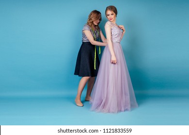 Beautiful Coordinator Seamstress Woman Button Up A Bride Wedding Dress In The Studio On A Blue Background . Tailor Dressmaker And Customer Prom