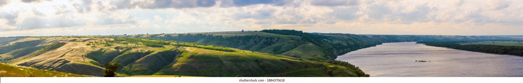 Beautiful countryside landscape. wide panoramic view of the river with a coastal meadow on the slope of a hilly coast, forests and a stormy dramatic dark sky with back sunlight from behind the clouds - Powered by Shutterstock