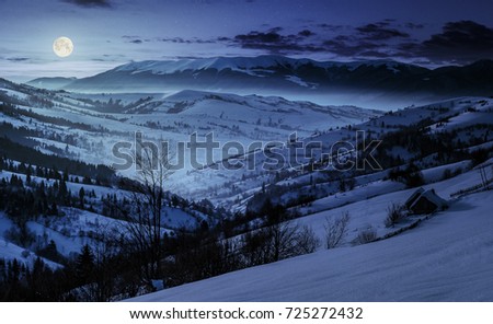beautiful countryside in mountains at night. village and rural fields on hillsides of valley covered with snow shine in full moon light
