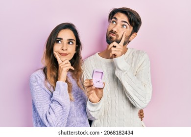 Beautiful couple of boyfriend and girlfriend holding engagement ring for proposal serious face thinking about question with hand on chin, thoughtful about confusing idea 