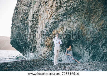Beautiful couple by the sea near the rocks, the girl bride sits, the groom stands beside her, happy and smiling
