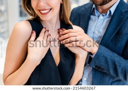 Beautiful couple enjoying in shopping at modern jewelry store. Young woman try it out gorgeous necklace and earrings. Selective focus on necklace with pendant. Fashion style and elegance concept.