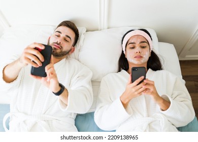 Beautiful Couple With Face Mask Wearing Robes And Relaxing Together In Bed While Texting On Their Smartphones