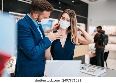 Beautiful couple with face protective masks enjoying in shopping at modern jewelry store. Young woman try it out gorgeous necklace and earrings. Fashion style and elegance concept. Pandemic lifestyle. - Powered by Shutterstock