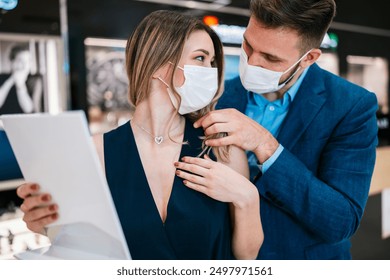 Beautiful couple with face protective masks enjoying in shopping at modern jewelry store. Young woman try it out gorgeous necklace and earrings. Fashion style and elegance concept. Pandemic lifestyle. - Powered by Shutterstock