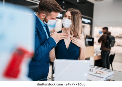 Beautiful couple with face protective masks enjoying in shopping at modern jewelry store. Young woman try it out gorgeous necklace and earrings. Fashion style and elegance concept. Pandemic lifestyle. - Powered by Shutterstock