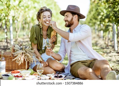 Beautiful couple having romantic breakfast with lots of tasty food and wine, sitting together on the picnic blanket at the vineyard on a sunny morning - Powered by Shutterstock