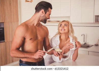 Beautiful couple in kitchen in the morning. Girl is drinking coffee while her shirtless boyfriend is making sandwiches, both are smiling - Powered by Shutterstock