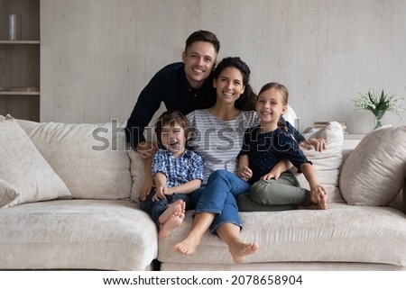 Beautiful couple with little children pose for photo. Preschool kids sit on sofa with Hispanic parents at home smile look at camera. Happy homeowners family, bank loan, medical insurance cover concept