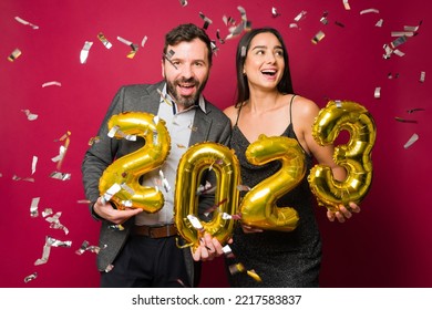 Beautiful couple looking excited celebrating new year's eve 2023 with balloons and confetti - Shutterstock ID 2217583837