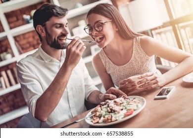 Beautiful couple in love is sitting in cafe. Young man is feeding his woman and smiling.