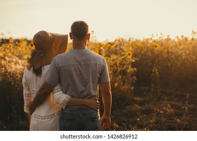 Beautiful Couple In Love Is Standing Back And Embracing, Looking At The Sunset Over A Field Of Sunflowers. Girl In A Hat And White Summer Dress, A Man In Jeans And A Tshirt. Summer Vacation Concept