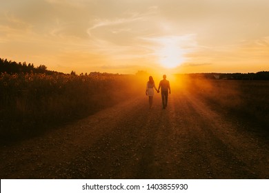 Beautiful couple in love is walking the road between the fields of sunflowers, summer sunset, bright orange light. In the background is a forest and rustic wooden houses. Concept of travel by car, eco