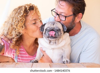 Beautiful Couple Of Man And Woman Kissing Their Old Pug Dog. Family And Love Concept. Wooden Table. Curly Hair For Her And Beard For Him