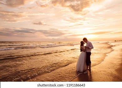 A beautiful couple of newlyweds, the bride and groom walking on the beach. Gorgeous sunset and sky. Wedding dresses, a white luxury dress for a girl. Family concept, honeymoon.