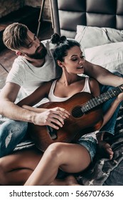 Beautiful Couple Playing Guitar. Handsome Young Bearded Man Teaching His Girlfriend To Play Guitar While Both Sitting In Bed At Home Together