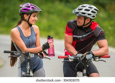 Beautiful couple riding on bicycles at the  road and looking to each other. Woman giving to man bottle of water.