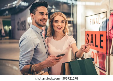 Beautiful couple with shopping bags is using a smartphone and smiling while doing shopping in the mall